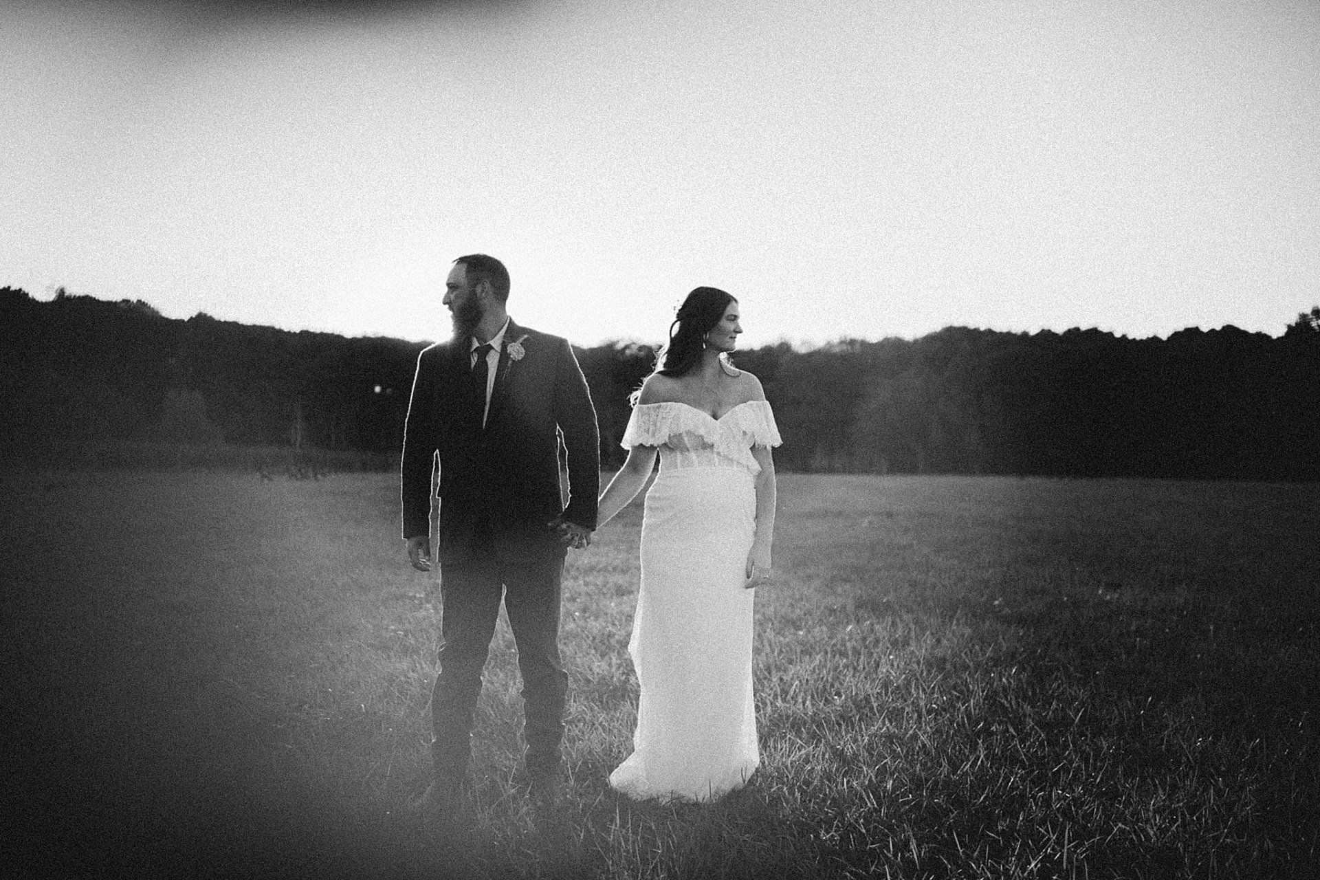 Couple in formal attire holding hands in a field at dusk. Sun shining behind them.