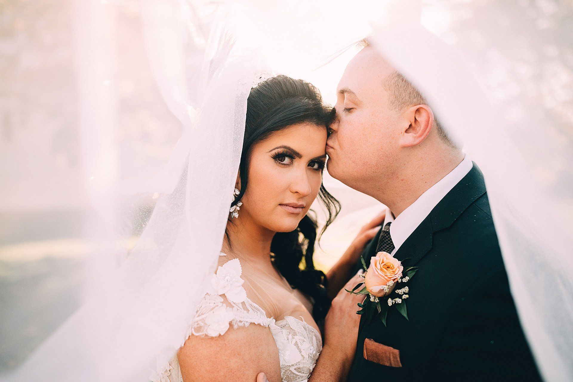 Bride and groom under a veil, groom kissing bride's forehead. Soft light, romantic wedding.