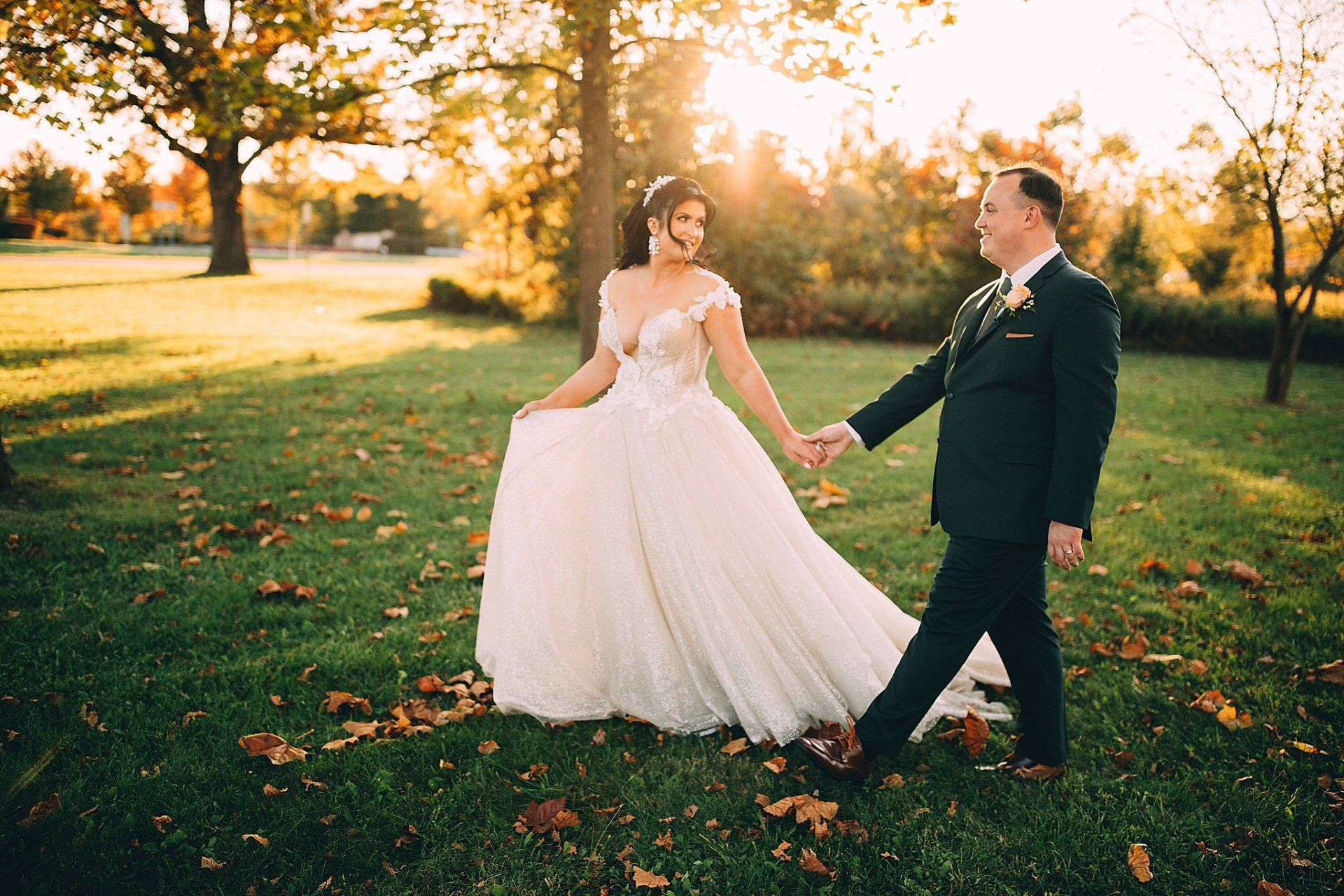 Bride and groom hold hands in a sunny field, bride in a white gown, groom in a dark suit, golden light.