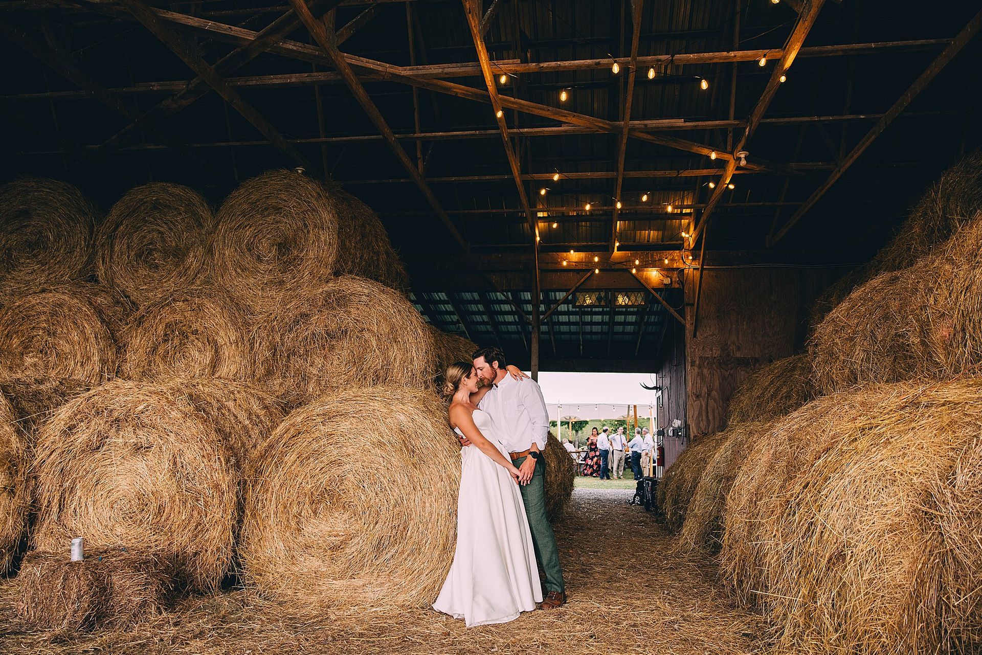 Couple embracing in a barn doorway, surrounded by hay bales, string lights overhead.