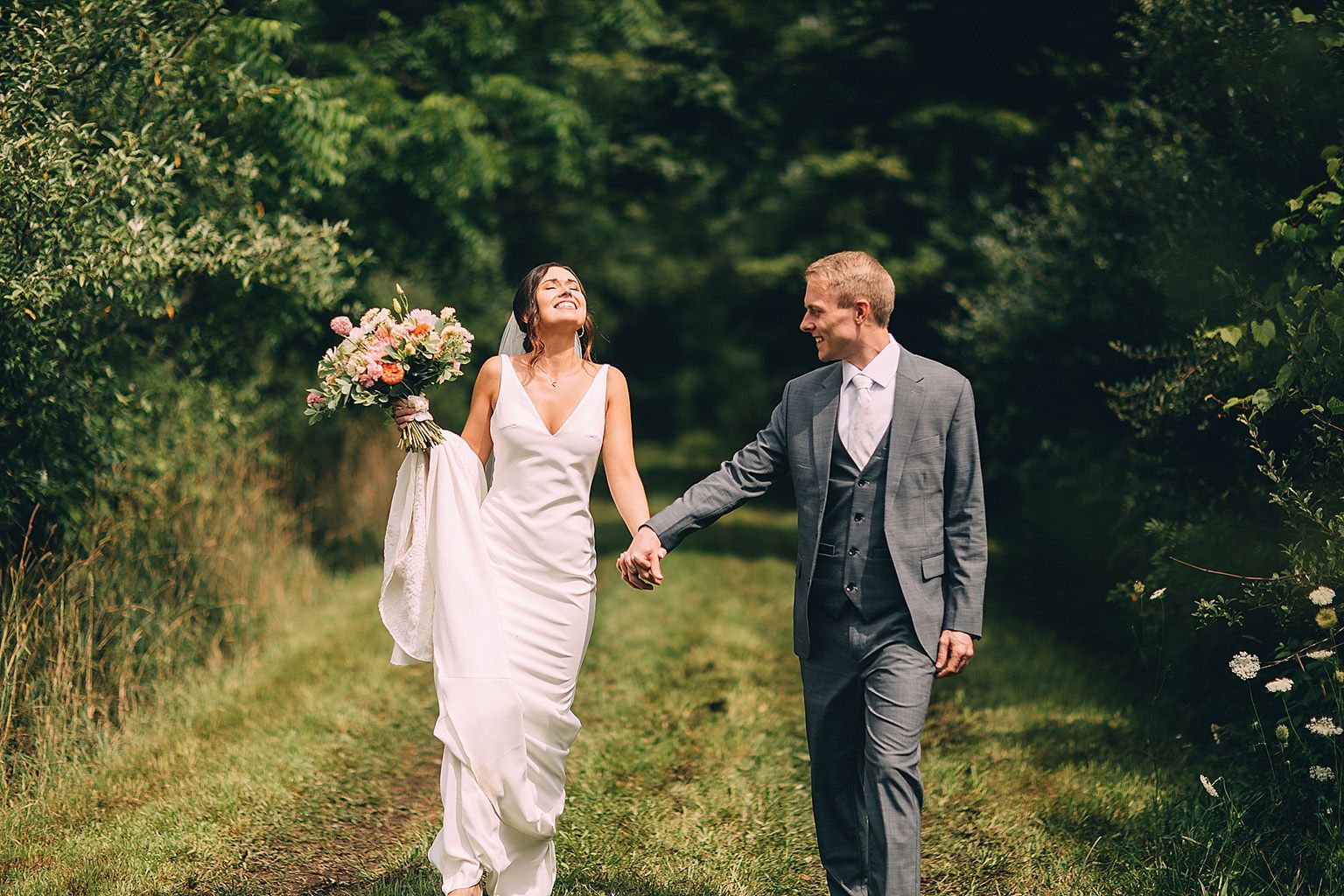 Bride and groom holding hands, smiling, walking on a path surrounded by greenery.