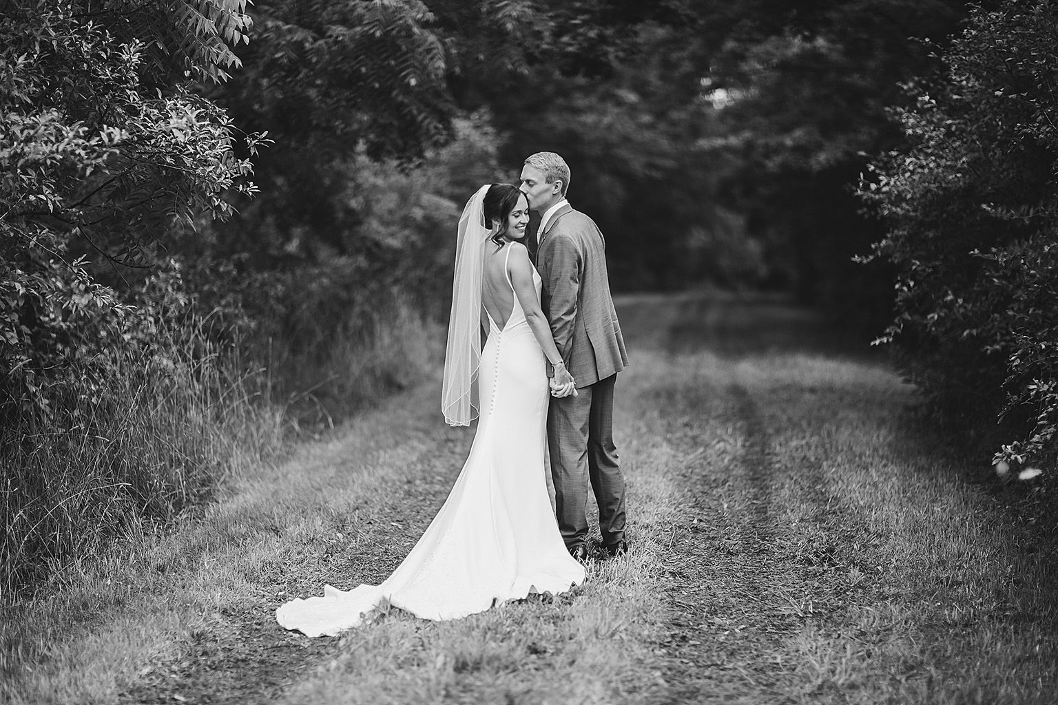 Wedding couple kissing on a grassy path surrounded by trees. Black and white.