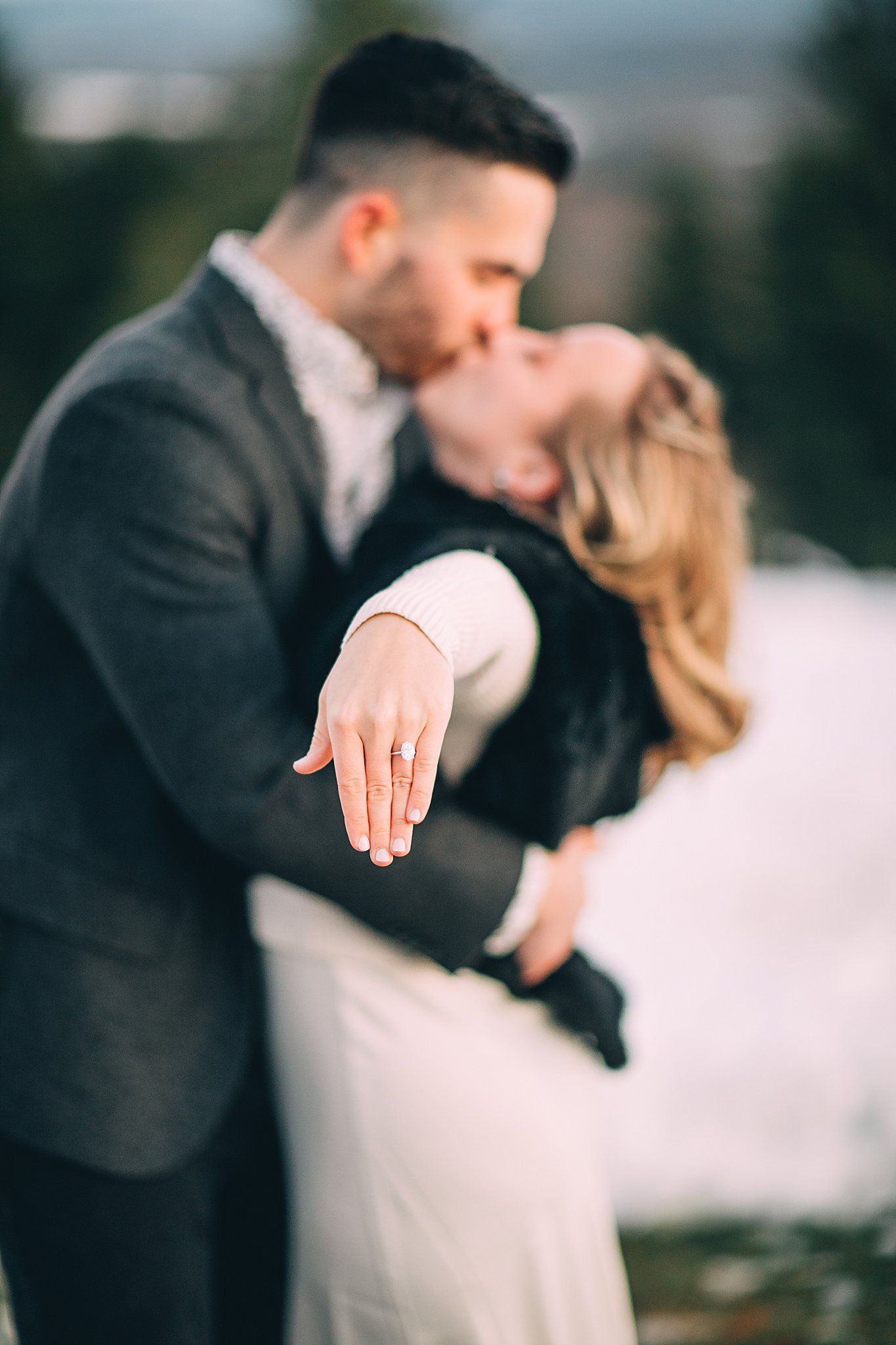 Couple kissing, woman showing off ring, outdoors, blurred background, snowy landscape.