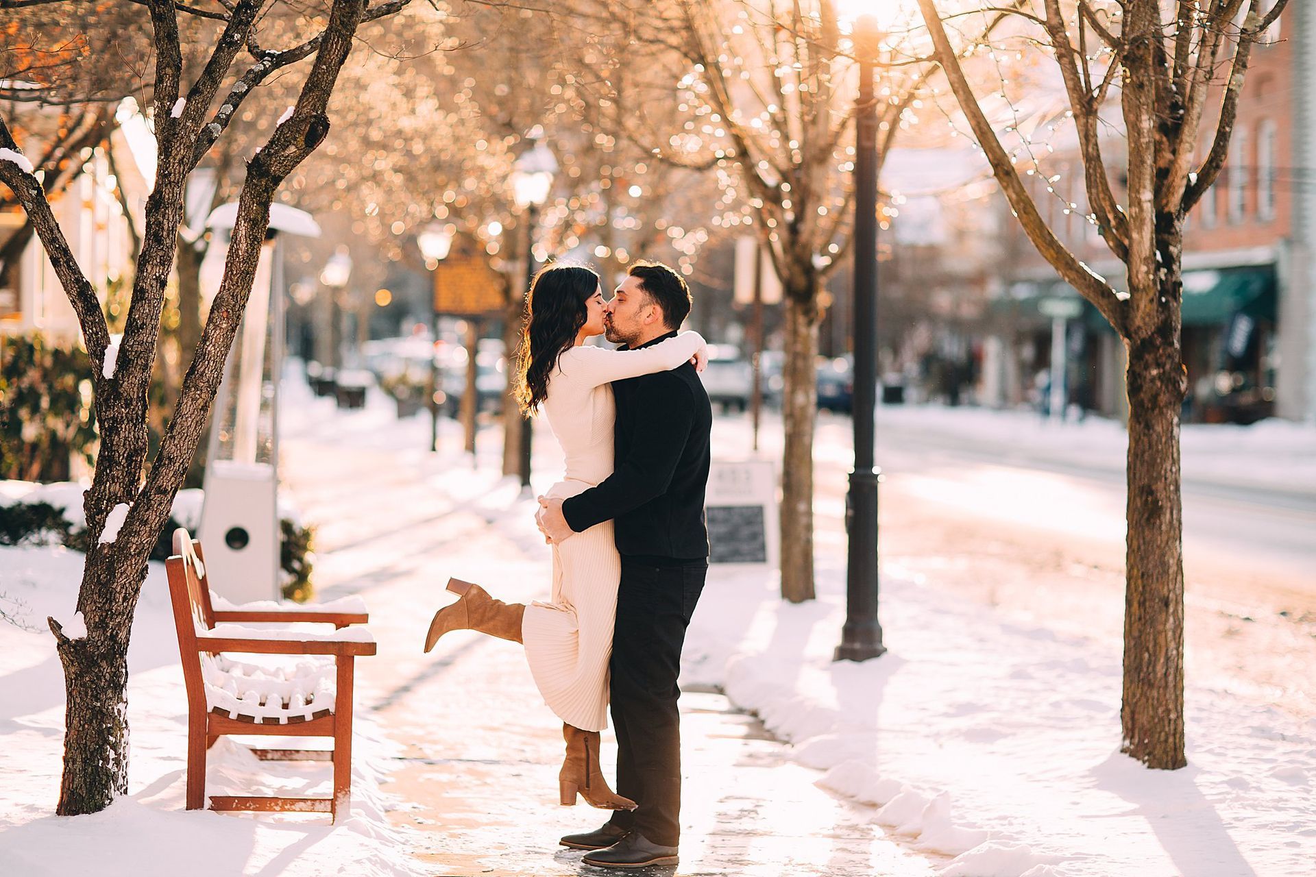 Couple embracing and kissing on a snowy street with a bench. Sunlight glows.