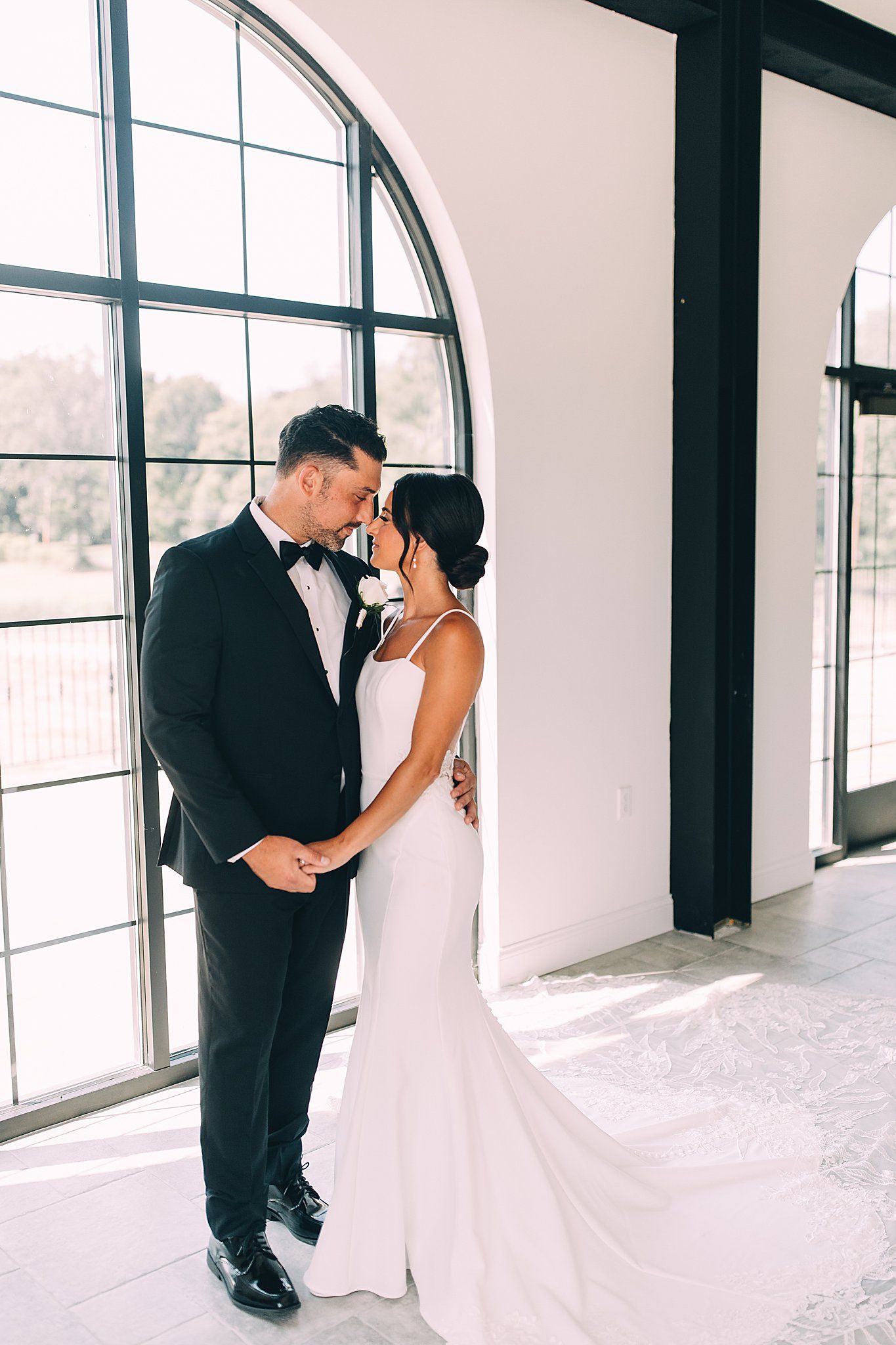Bride and groom holding hands, foreheads touching, in front of large arched window.