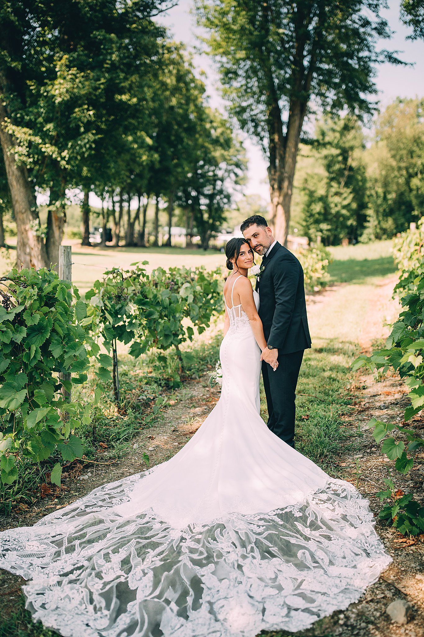 Couple in wedding attire posing in a vineyard, holding hands, dress with long train.
