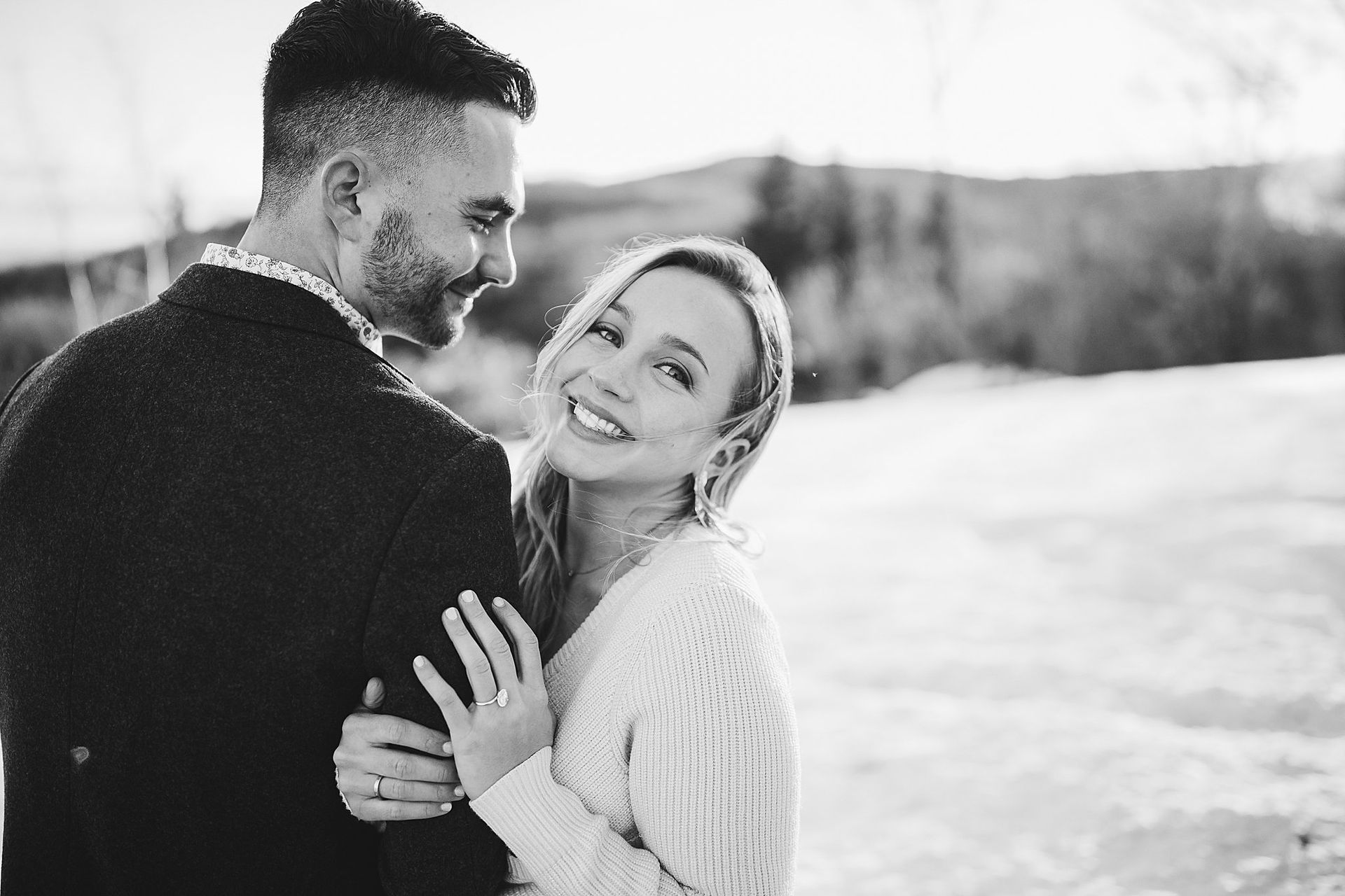 A smiling woman in a sweater leans on a man, both smiling, in a snowy outdoor setting.