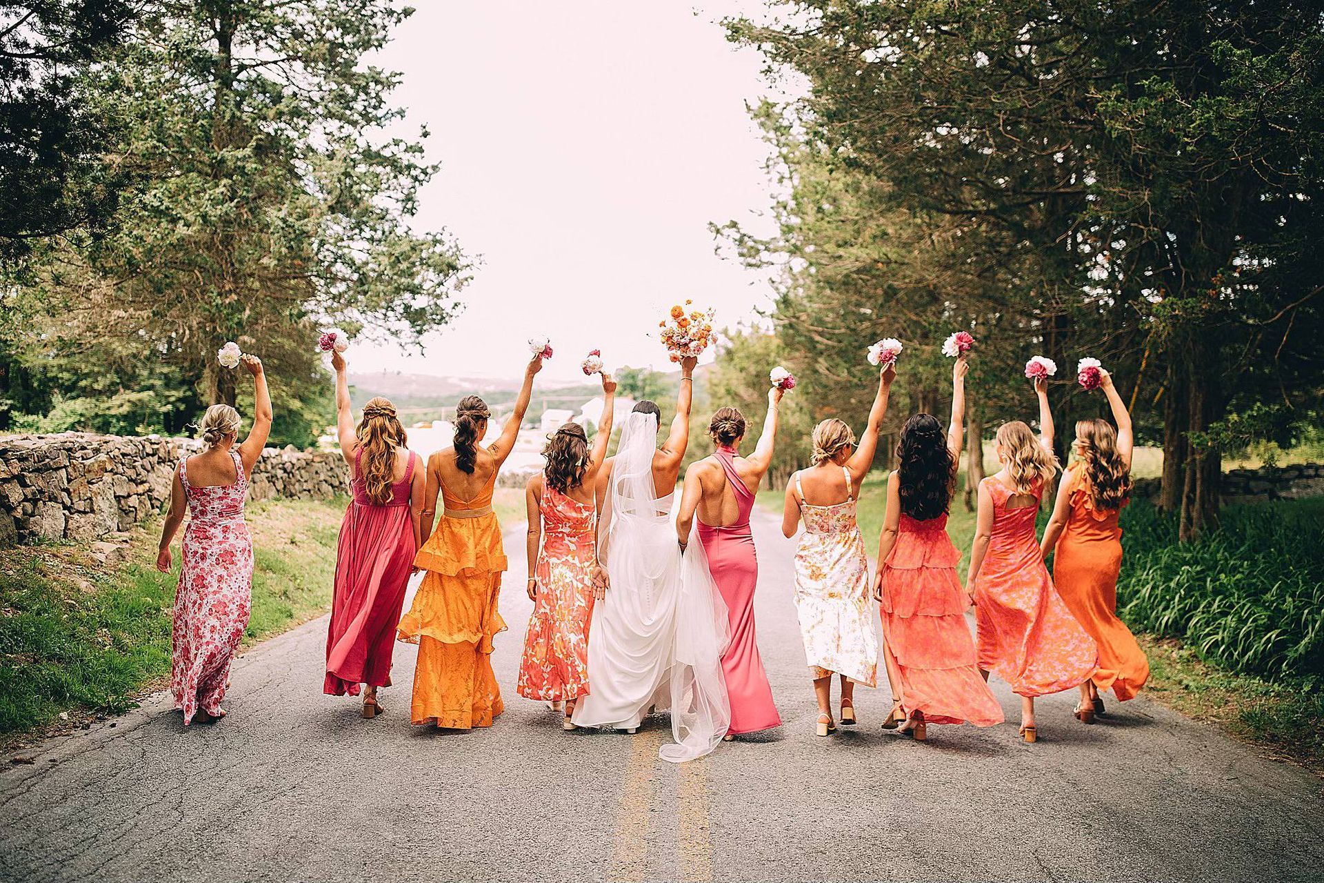 Bridesmaids in colorful dresses raise bouquets, walking away from the camera on a gravel road.