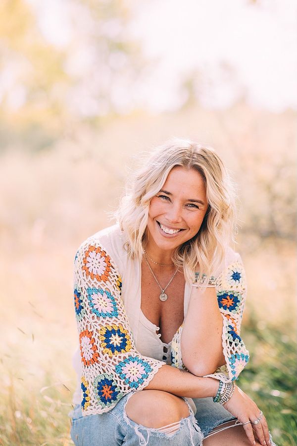 Woman smiles, seated in tall grass, wearing crochet top and ripped jeans.
