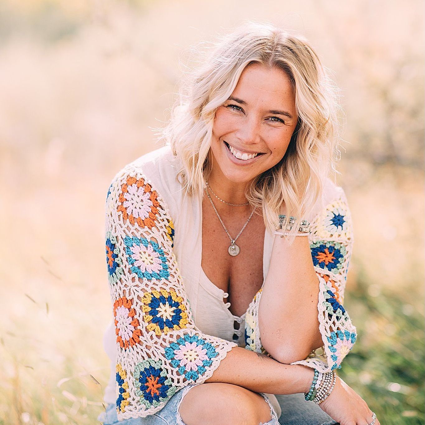 Blond woman smiling, wearing a colorful crocheted top, sitting outdoors in a field, sunny.