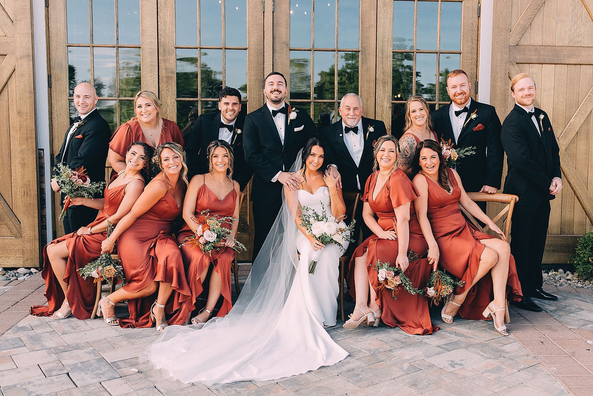 Wedding party in front of wooden doors. Bride in white gown, bridesmaids in rust dresses, groomsmen in black tuxedos.