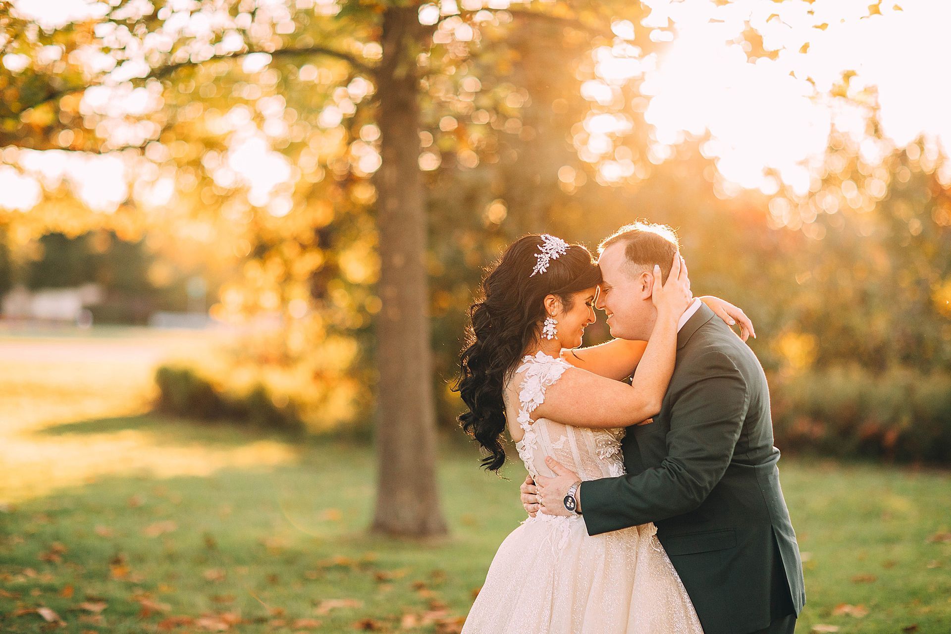 Couple embracing outdoors, sunlight filtering through trees. Bride in white gown, groom in suit.