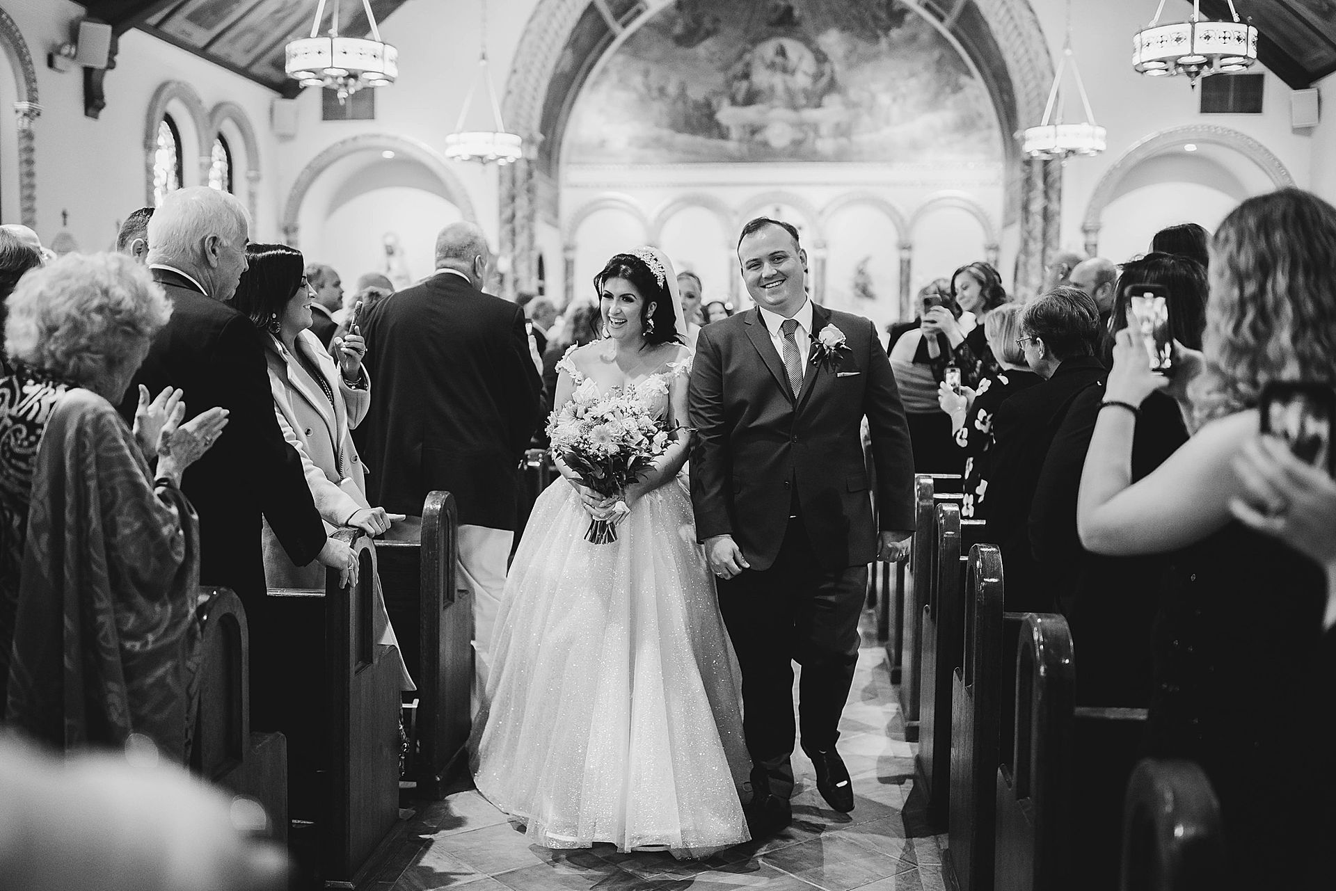 Newlyweds walk down the aisle of a church, smiling, as guests clap and take photos.