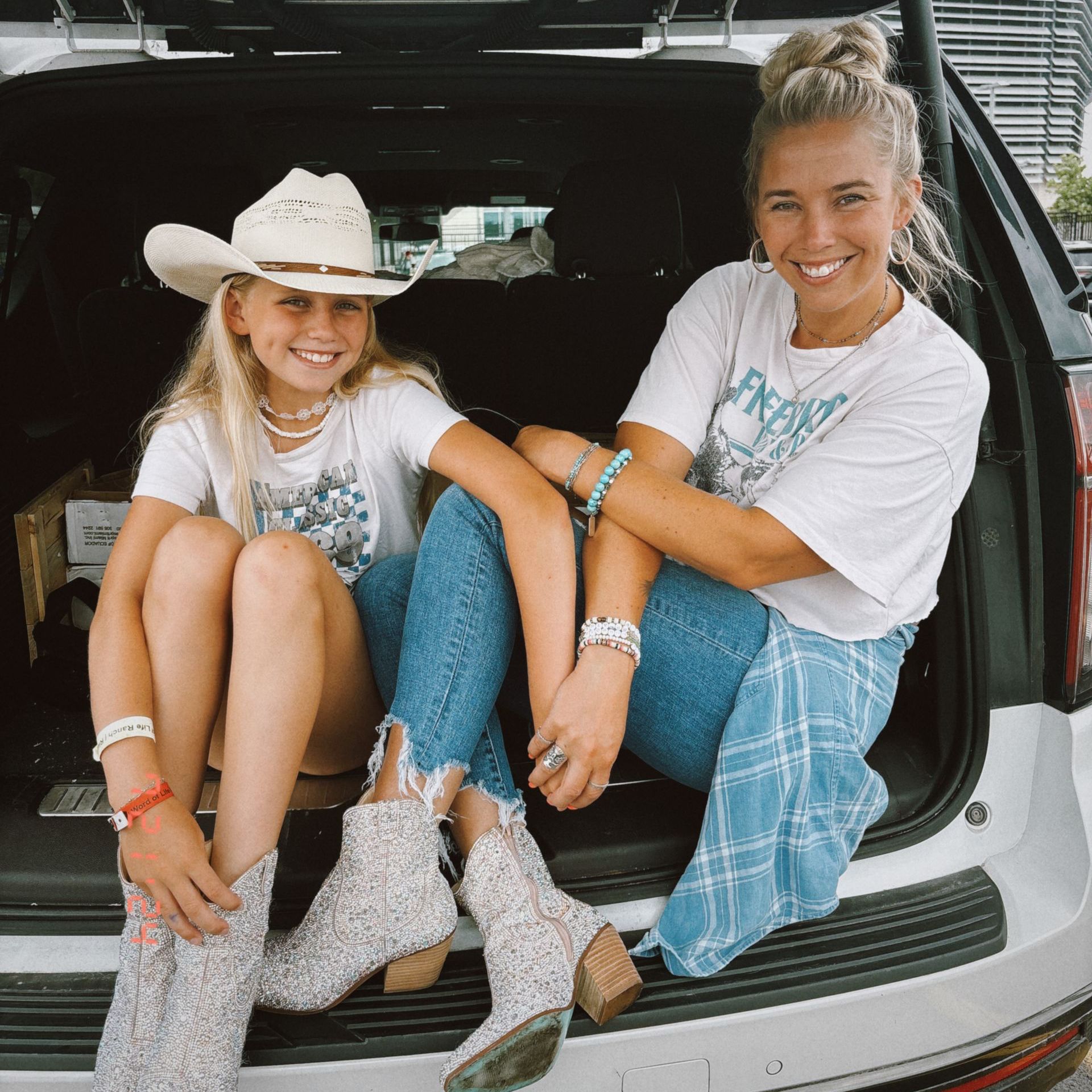 Two women in a car trunk, smiling. One in a cowboy hat and boots; other in a bun, jeans.