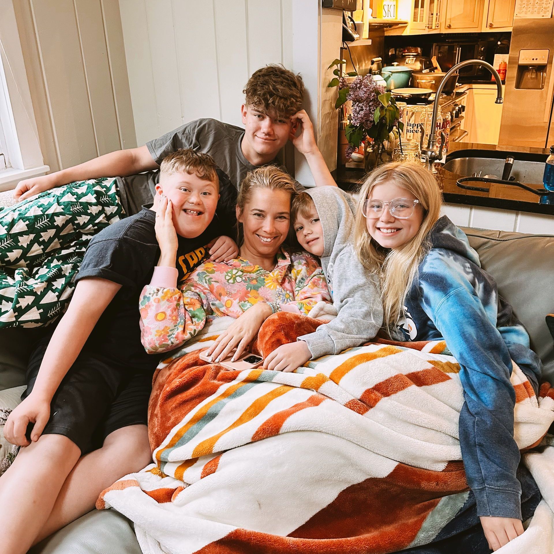 A woman sits on a couch, surrounded by five children, all smiling. They are covered by blankets.
