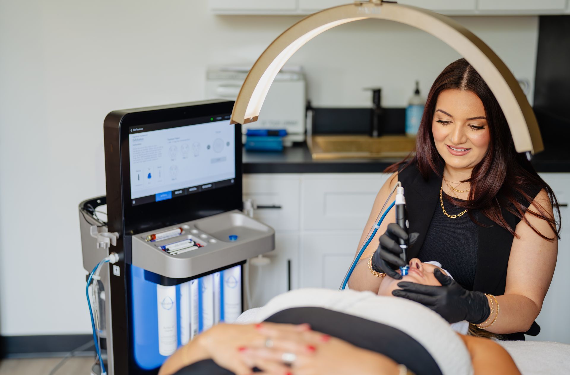 Woman performing facial treatment on a client in a clinic. She uses a device with a screen and light.