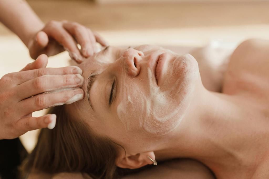 Woman receiving a facial treatment, lying down with eyes closed, hands on her face.