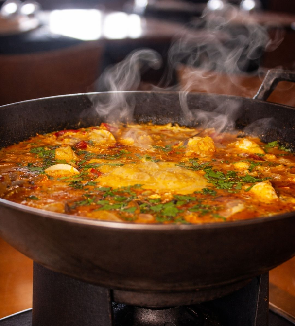 Tigela fumegante de sopa ou guisado vermelho-alaranjado com ervas e bolinhos em uma panela preta.