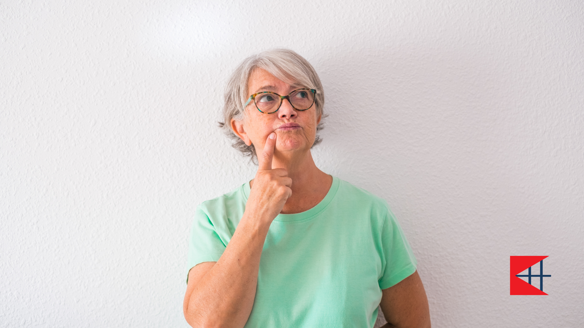 Woman in glasses, light green shirt, touching chin, looking upward, thinking, against white wall.