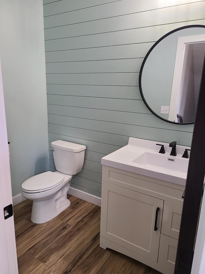 Small bathroom with white toilet, sink vanity, round mirror, pale green shiplap wall, and wood-look floor