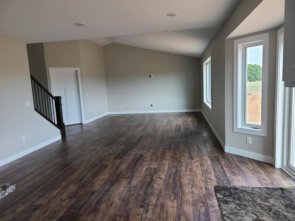 Empty living room with dark wood floors, gray walls, staircase, and large windows.
