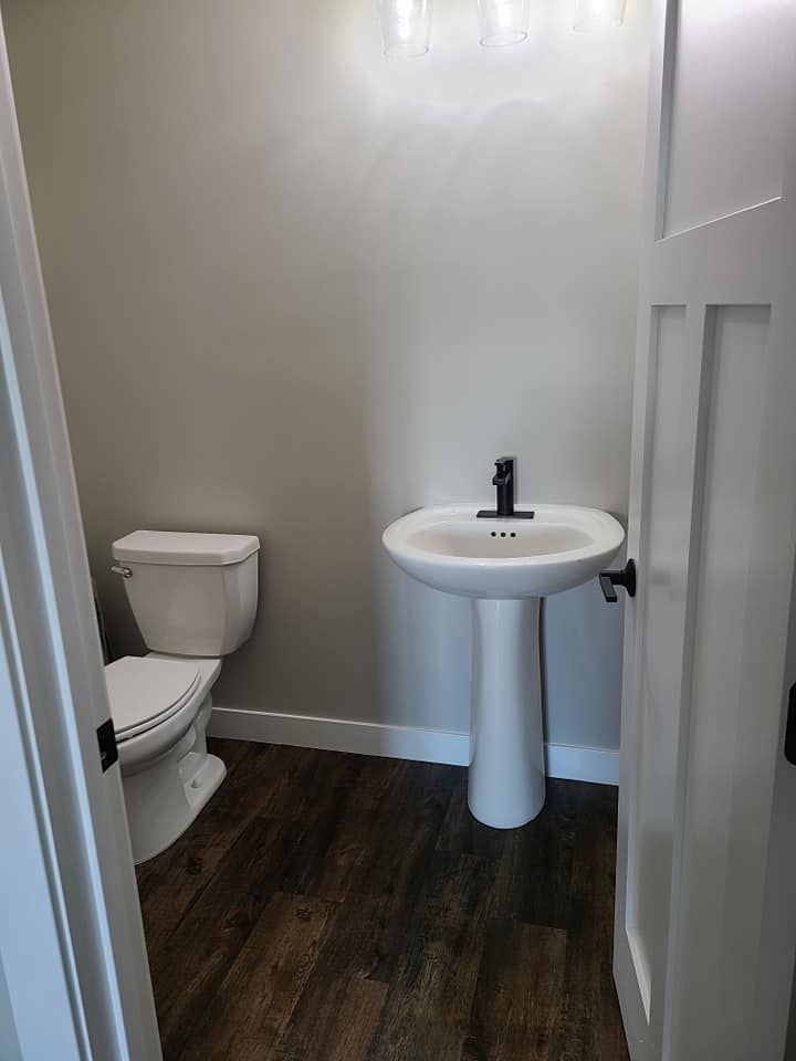 Small bathroom with toilet, pedestal sink, white walls, and dark wood-look floor.