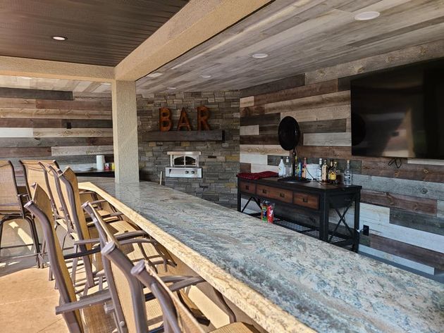Covered outdoor bar with stools, stone countertop, TV, grill, and wood-paneled ceiling.