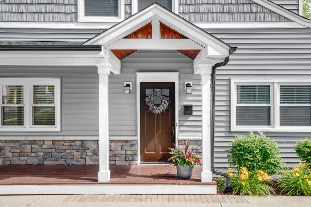 Front entrance of a gray house with white trim, wood door, potted plants, and a small porch