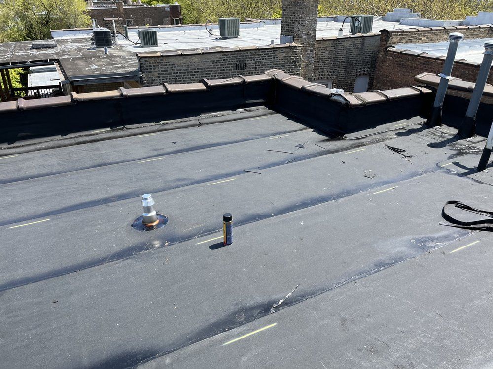 Flat black rooftop with vents, pipes, and a stone parapet under bright daylight