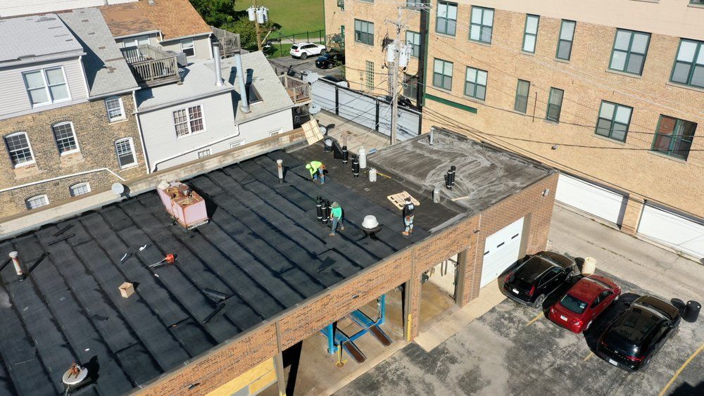 Workers on a flat black rooftop beside brick buildings and parked cars in a courtyard.