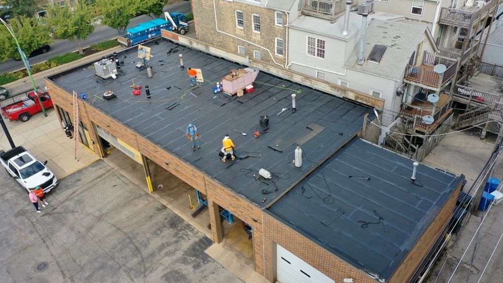 Aerial view of workers installing a dark roof on a flat-roofed building beside a street.