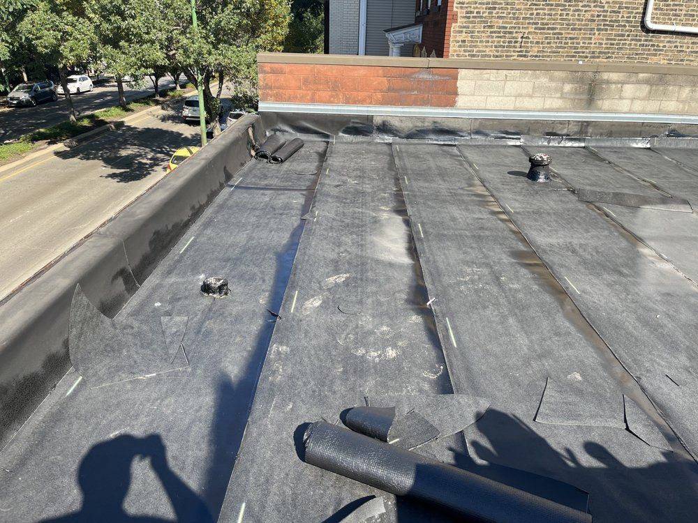Flat rooftop with black rolled roofing, roof vents, and a brick parapet beside a tree-lined street.