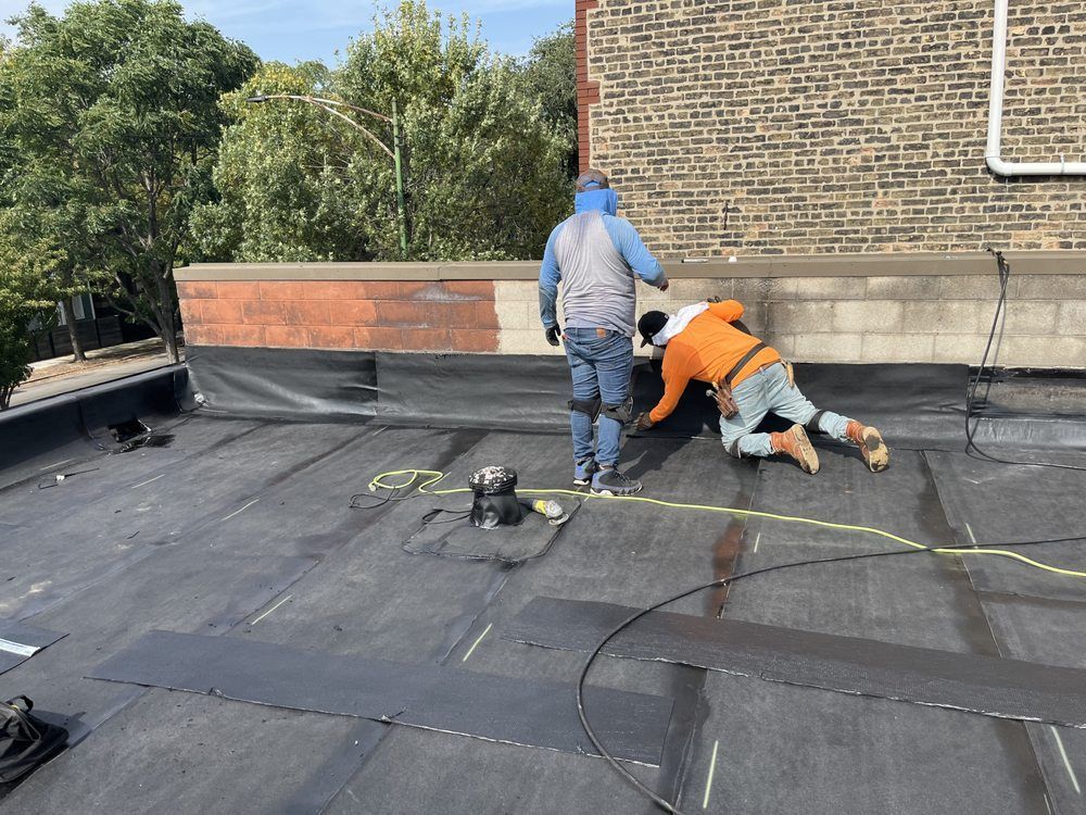 Workers repairing a flat roof with roofing tools near a brick wall and trees in the background