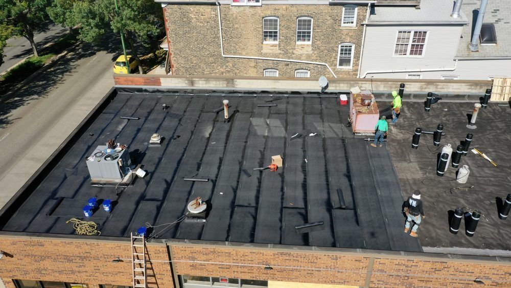 Workers installing black roofing on a flat rooftop beside a tan brick building.
