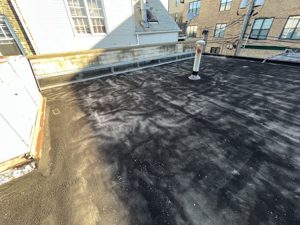 Flat black rooftop with a vent pipe and sunlight casting shadows beside brick buildings.
