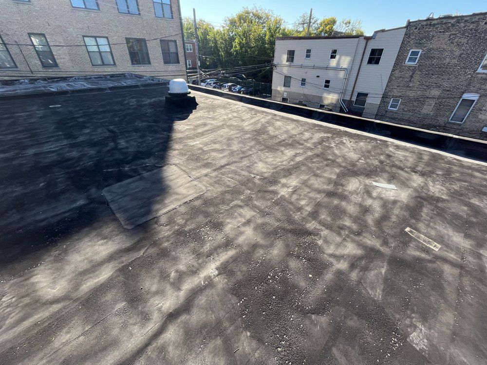 Flat black rooftop with a white vent, bordered by brick apartment buildings in sunlight.