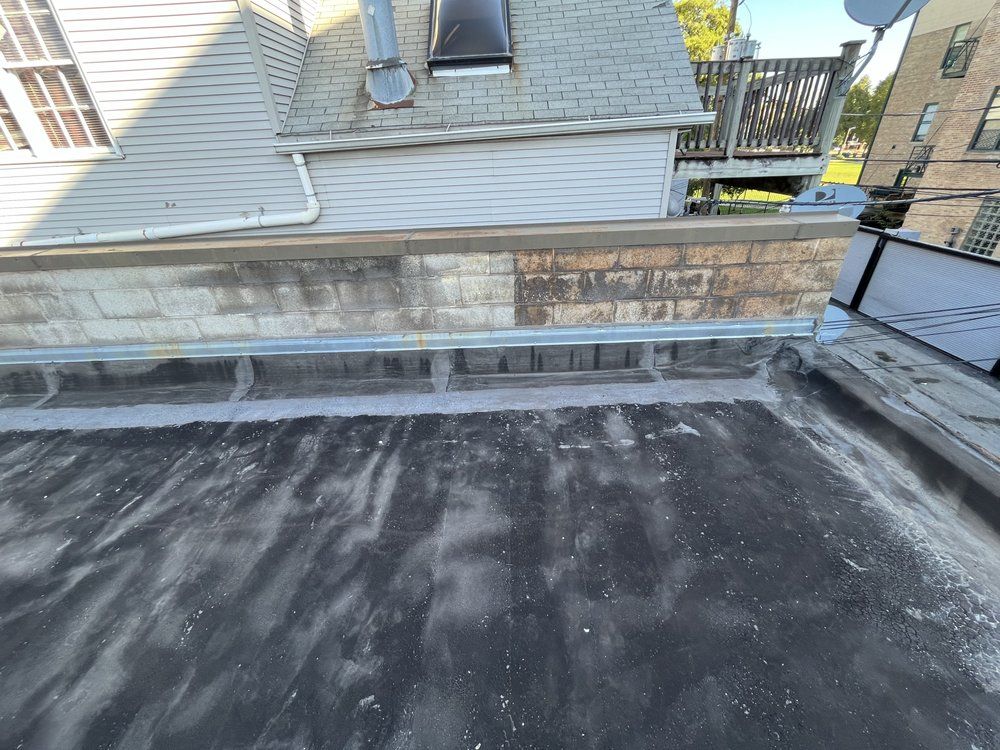 Rooftop with dark membrane surface and a low stone parapet, viewed beside neighboring houses.