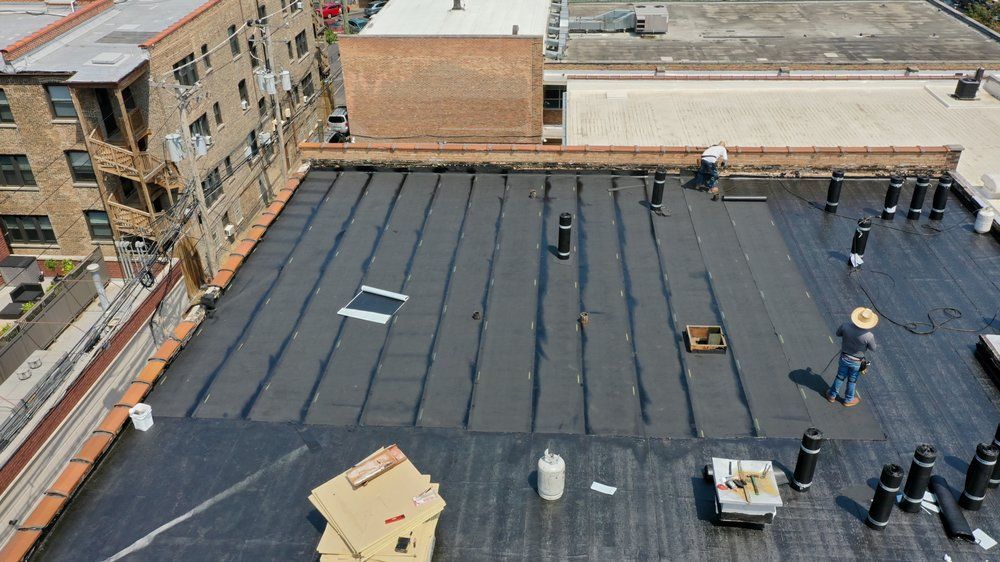 Rooftop with black membrane, HVAC units, vents, and adjacent brick buildings under bright daylight