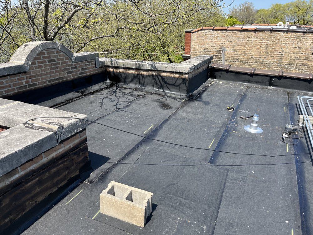Flat black rooftop with brick chimney structures and a small cinder block, trees in the background