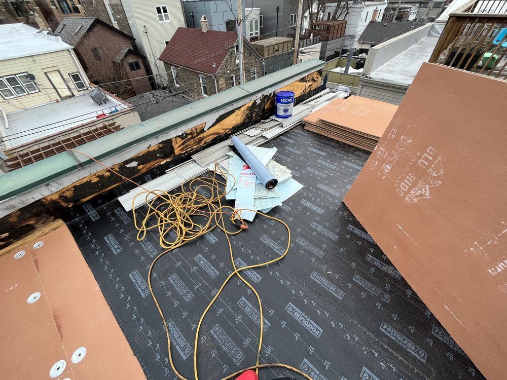 Rooftop under construction with black roofing underlayment, coiled cables, and scattered tools near a brick parapet.