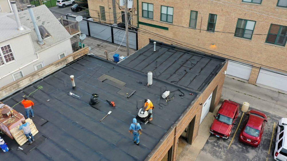 Workers on a flat rooftop installing a black roof membrane beside neighboring buildings and parked cars.