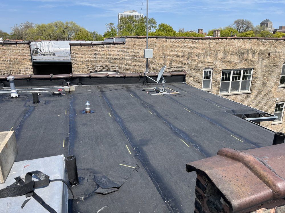 Flat rooftop with dark membrane, vents, and a brick parapet against a stone building and blue sky.