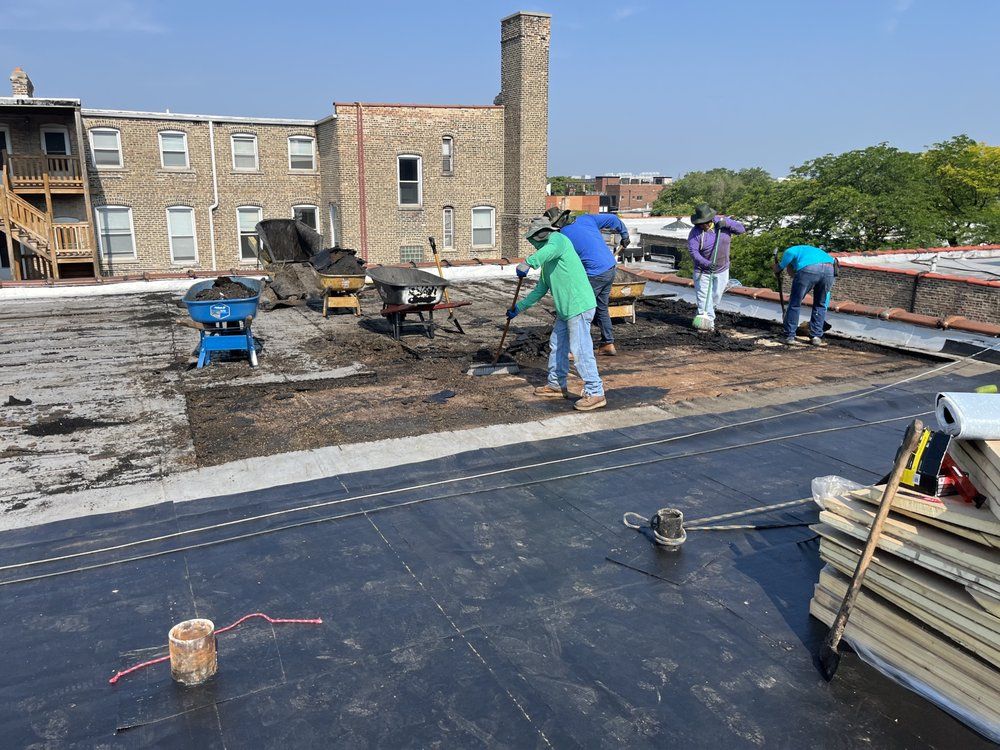 Workers on a flat rooftop laying roofing materials near a brick building under a clear sky