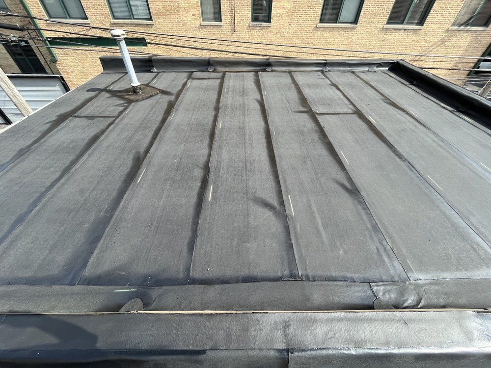 Flat black roof with seams and vent pipes against a brick building backdrop