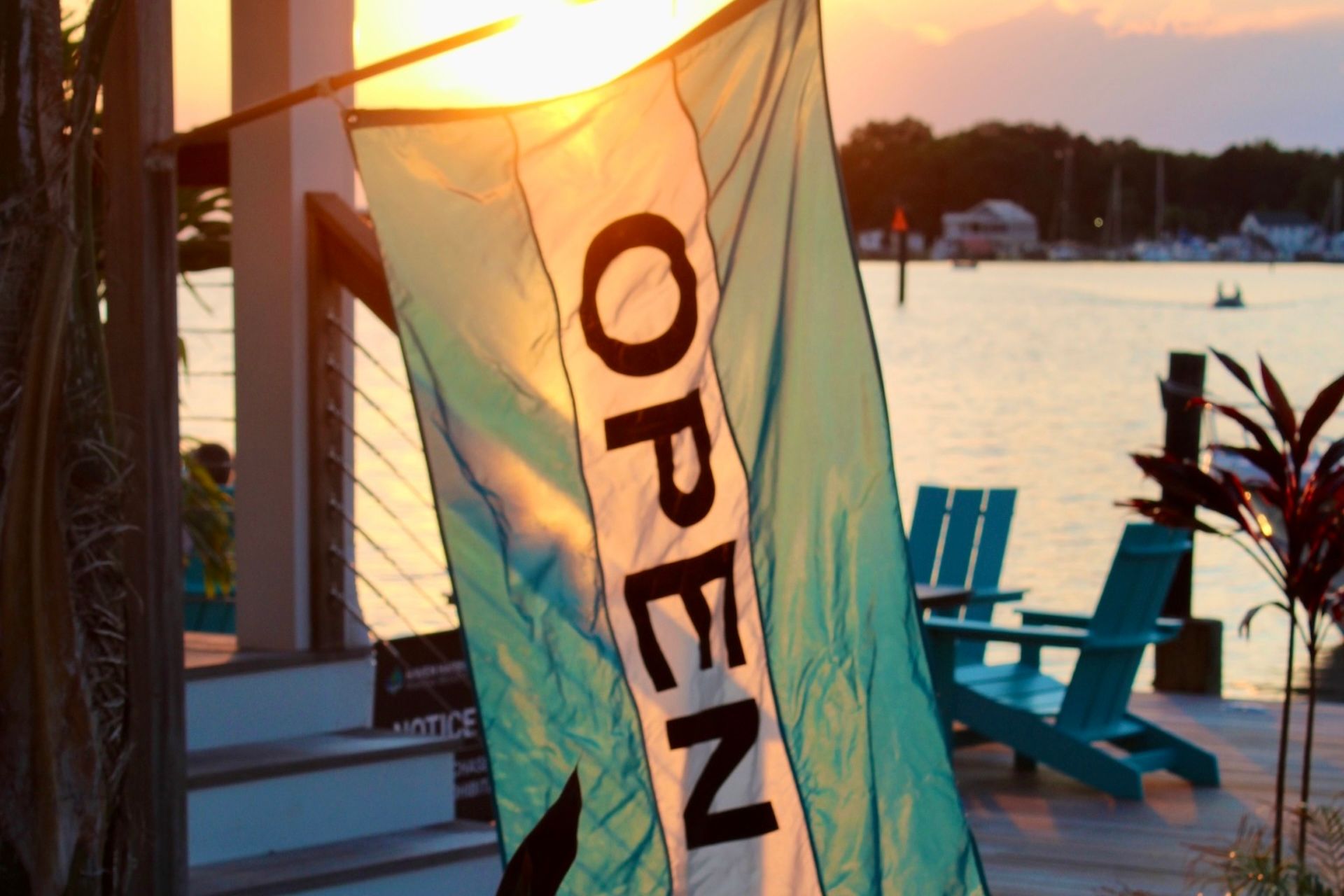 Sunlit waterfront dock bar with an open flag and blue chairs at sunset.