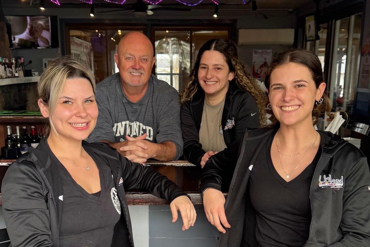 Group of three women and one man pose for photo in an empty bar room