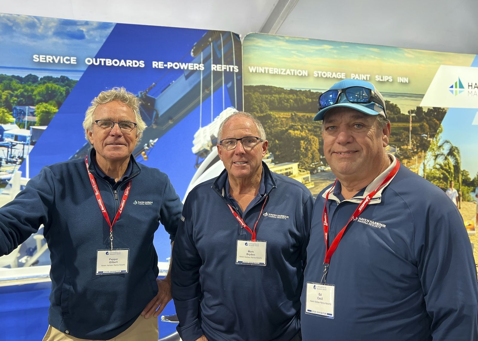 Three men in navy shirts and red lanyards stand smiling at a boat show booth.