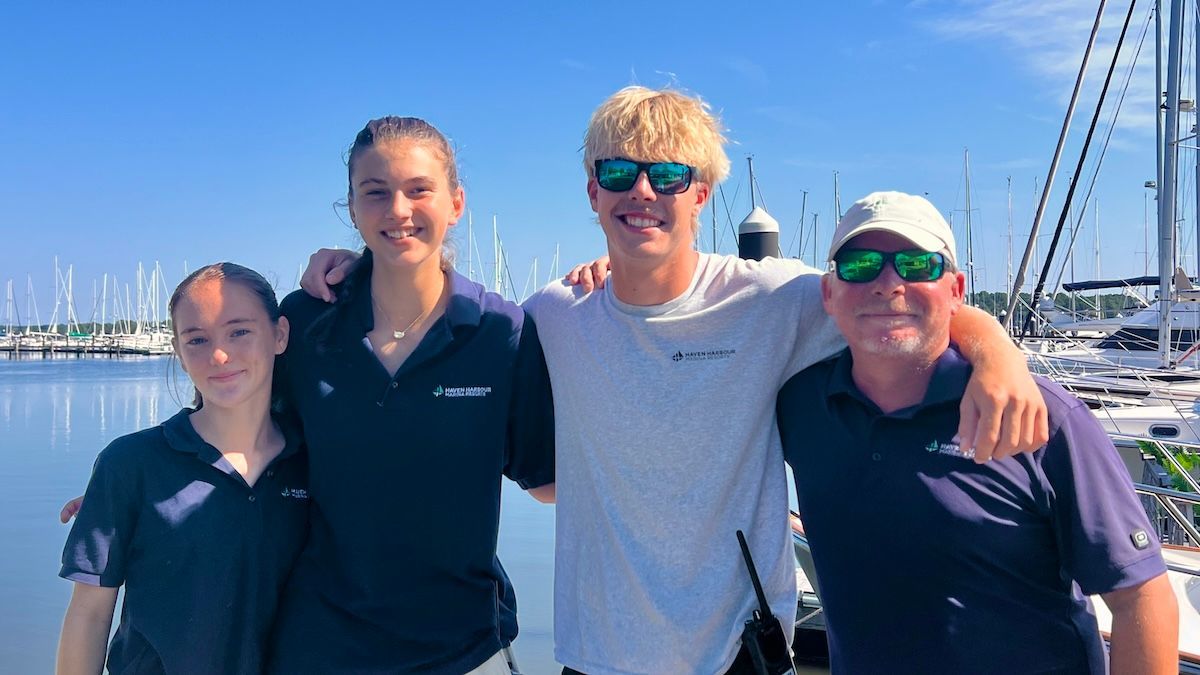 Four people smiling on a sailboat dock, with blue water and sailboat masts in the background.