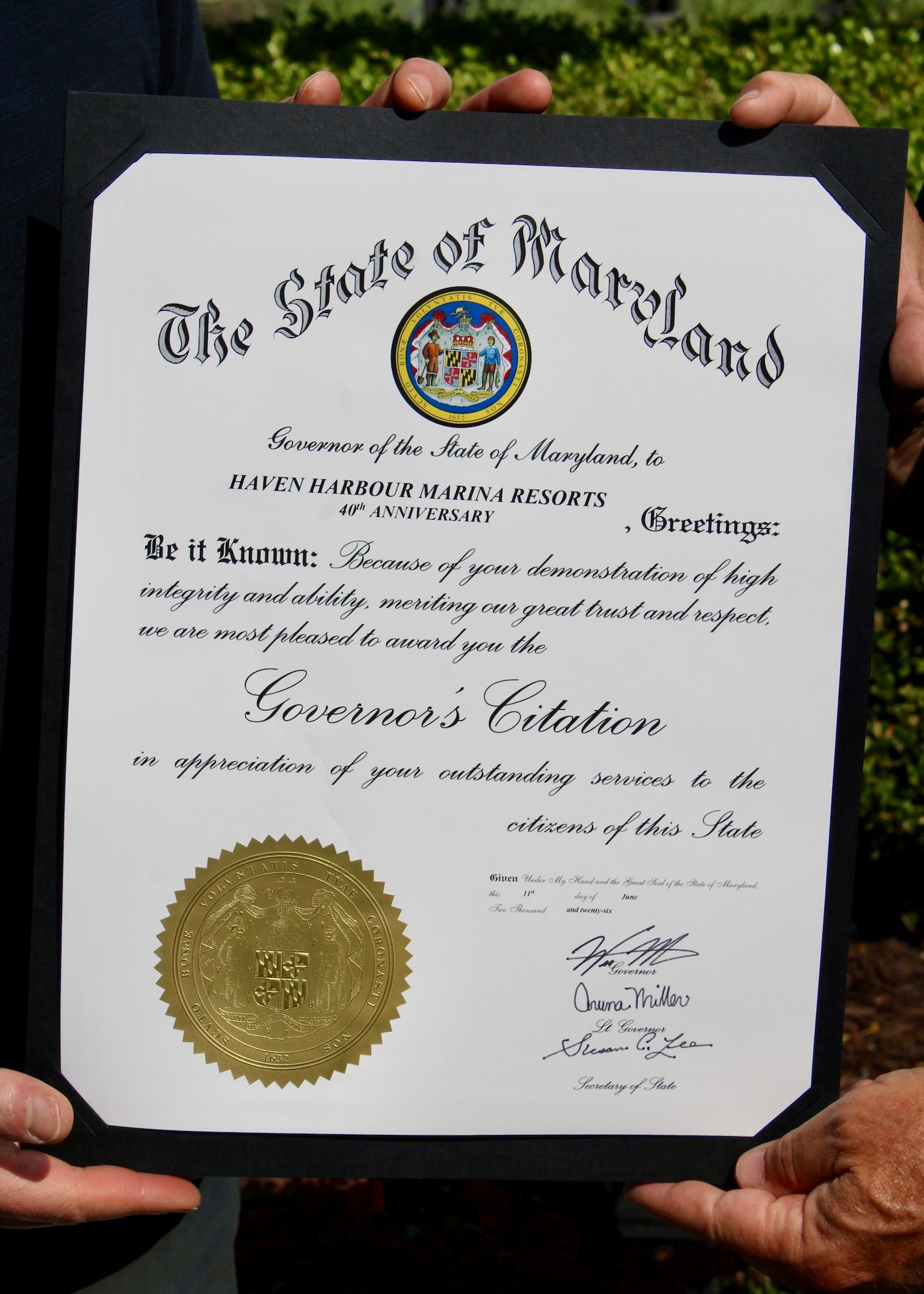 Maryland governor’s citation on white paper with gold seal, held up by two hands outdoors.
