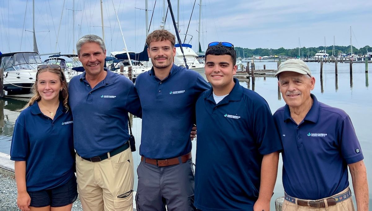 Five people in navy polo shirts stand smiling on a dock with sailboats and water behind them.